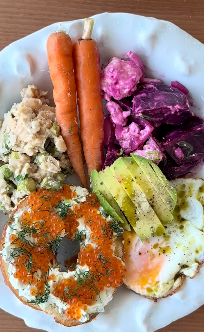 Plate with bagel topped with cream cheese and roe, sliced avocado, poached egg, beet salad, mixed potato salad, and two carrots