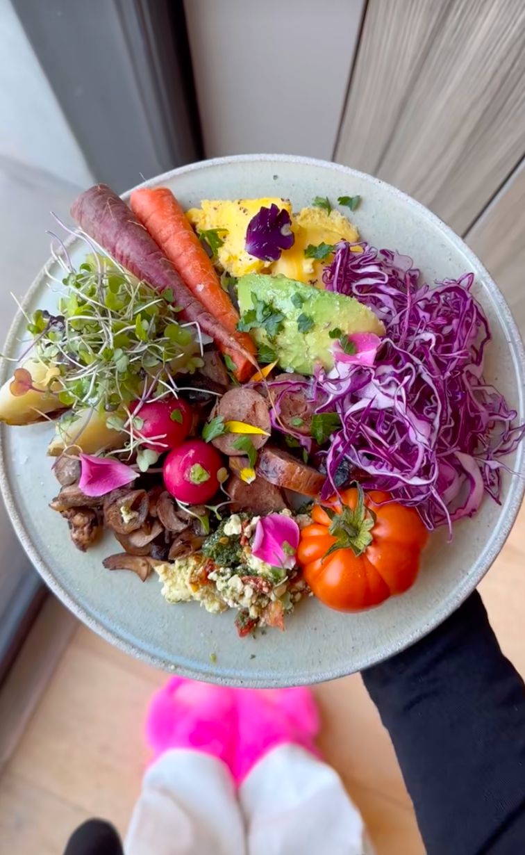 A vibrant, artfully arranged vegetarian dish featuring assorted fresh vegetables, greens, a slice of salami, and edible flowers on a round plate