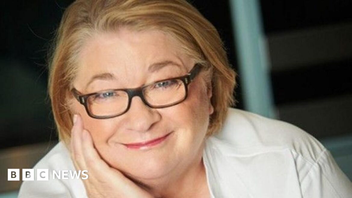 TV chef Rosemary Shrager starts 450-mile charity bike challenge Rosemary Shrager in jeans, a bright yellow high vis jacket and a bike helmet. She is standing on a road next to a bike.