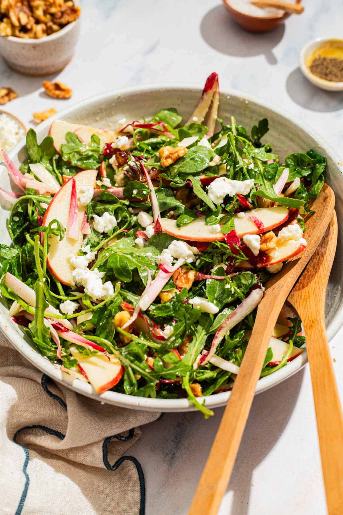 Apple walnut salad in a serving bowl with wooden serving utensils surrounded by bowls of salt and pepper and a cloth napkin.