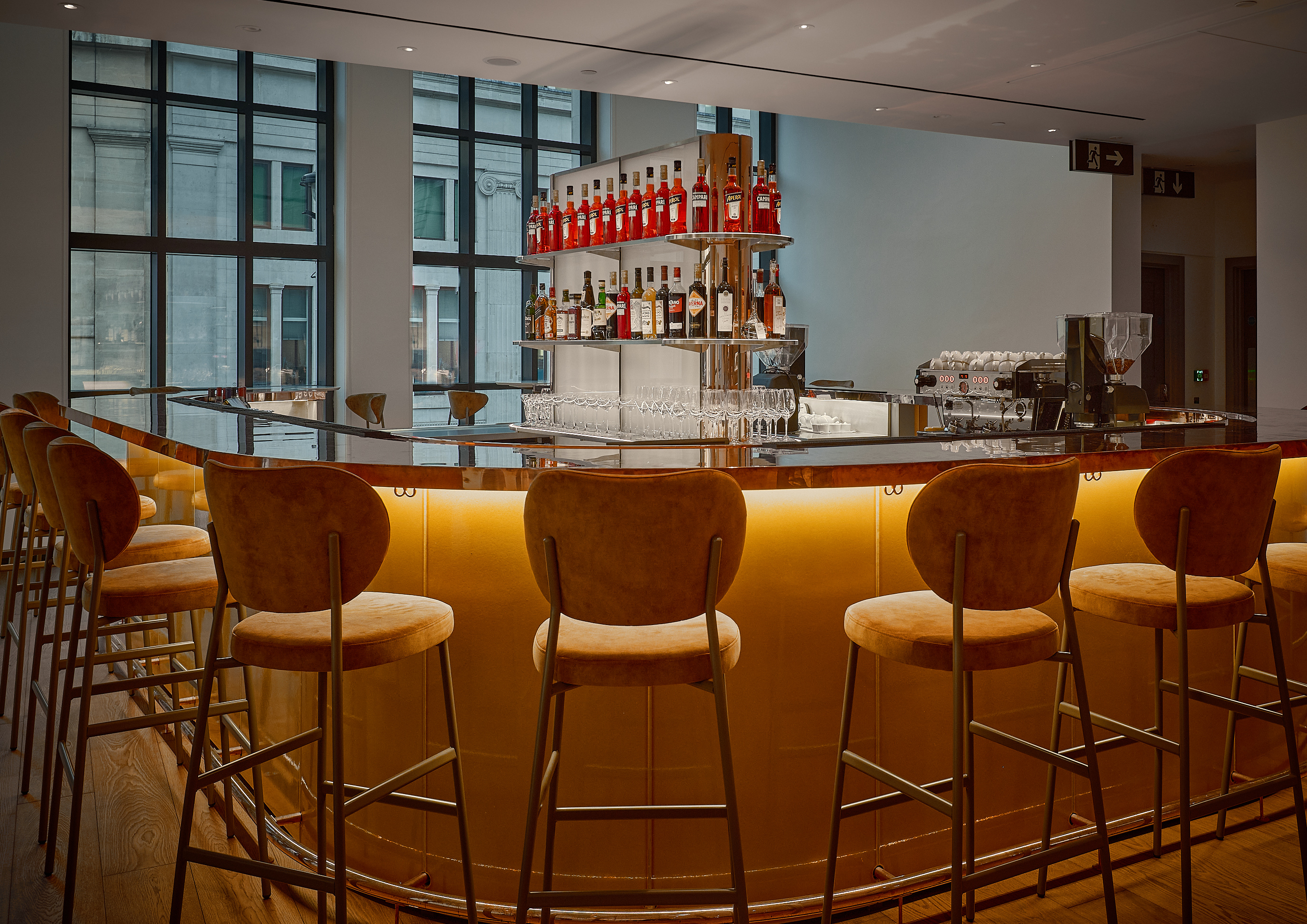A dazzling cocktail bar features a sleek bronze countertop, encircled by velvety brown stools, and a central cabinette filled with all sorts of liqueurs.