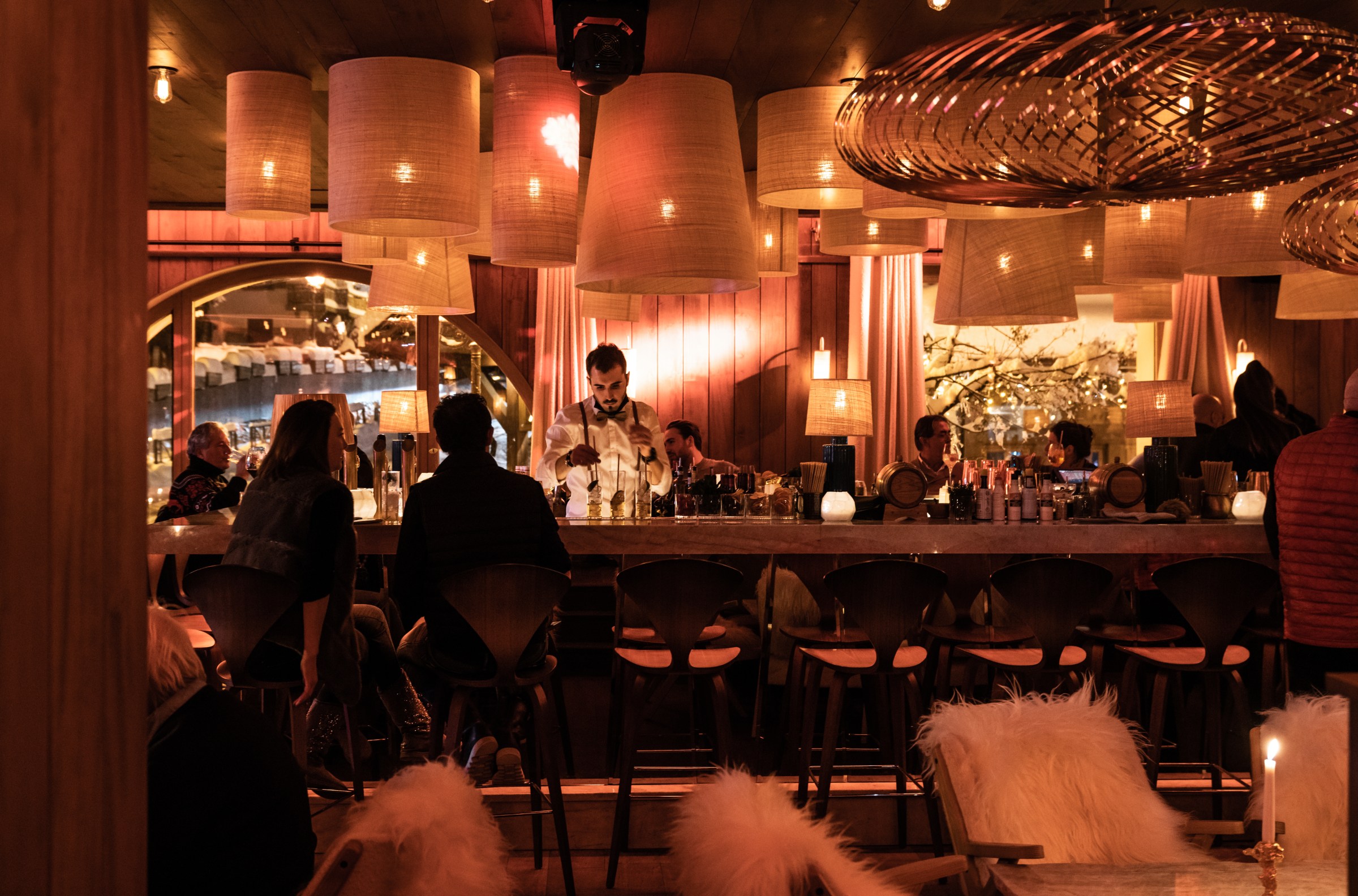 A dark bar interior with a bartender working behind the bar, customers at bar stools, and soft lighting from a variety of high-design lights