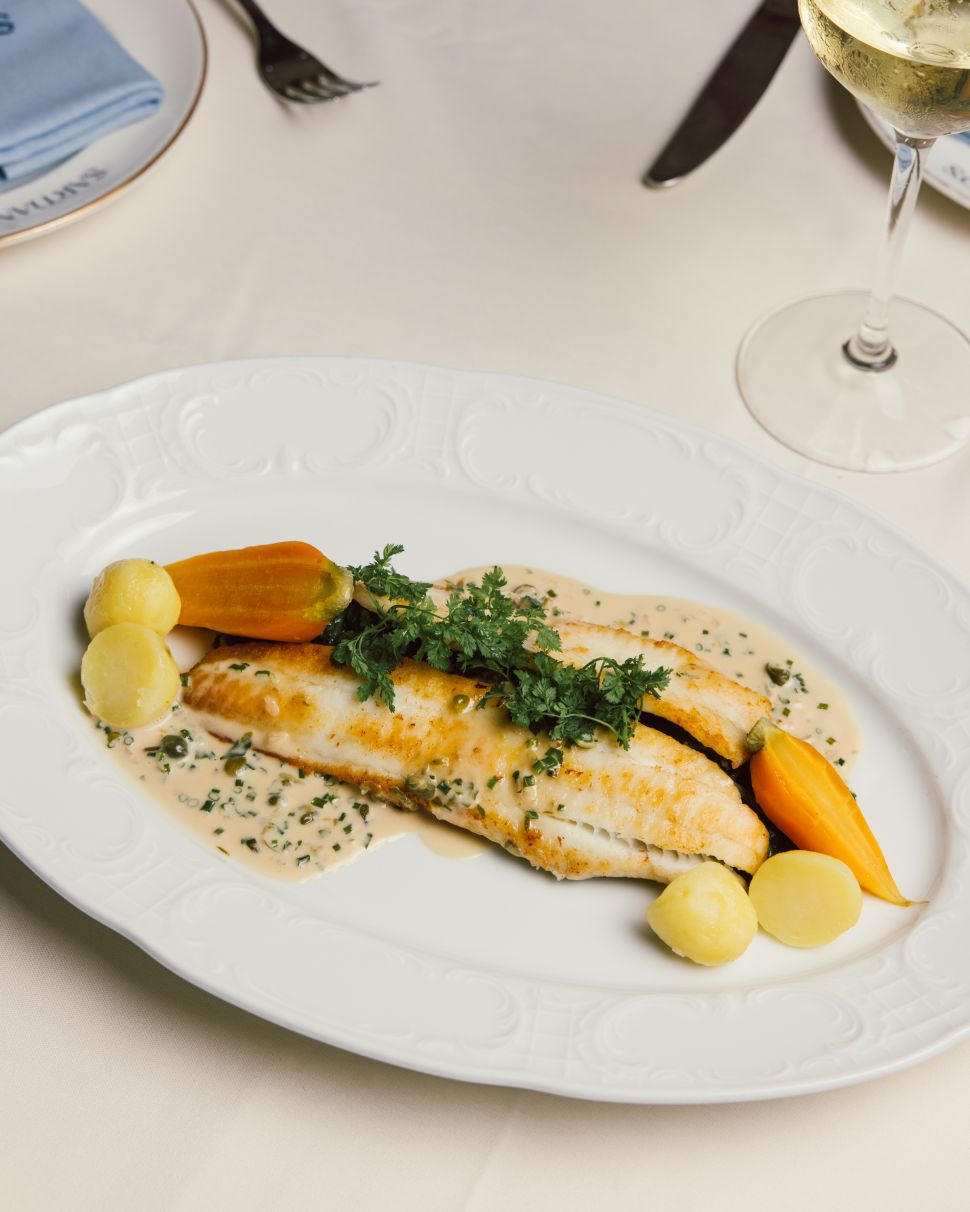 A refined plate of Dover sole piccata, elegantly garnished with fresh vegetables and herbs, accompanied by a creamy sauce, placed on a white plate.
