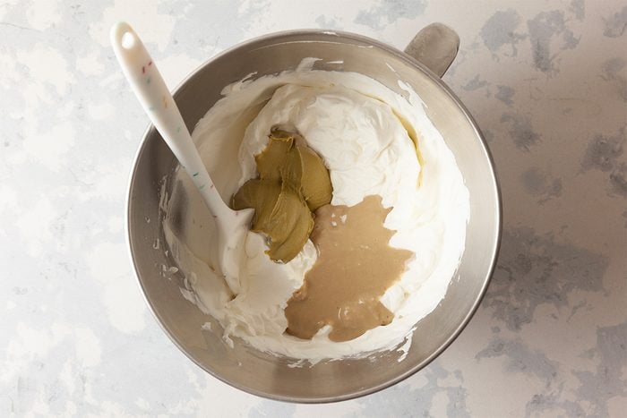 A metal mixing bowl containing white creamy mixture, pickle slices, and a light brown liquid, with a white spatula resting inside the bowl on a light-colored surface.