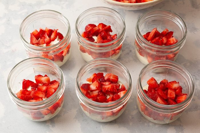 Six glass jars arranged in two rows, each filled with a layer of creamy dessert topped with diced fresh strawberries, set on a light-colored surface.