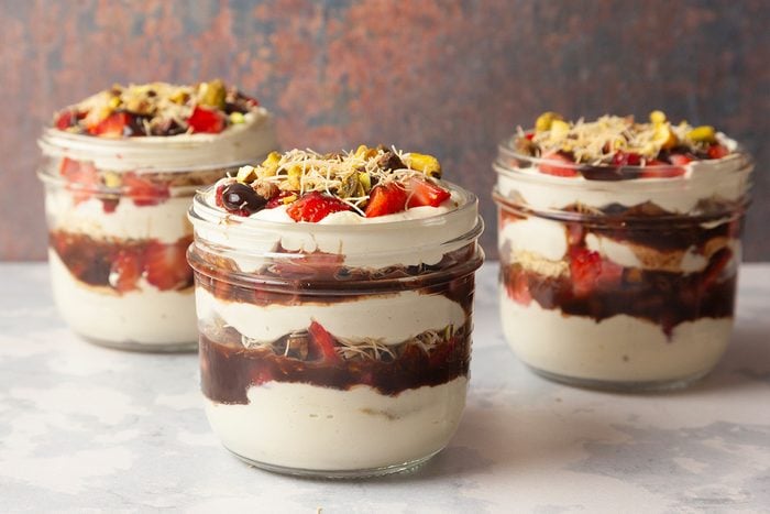 Three glass jars filled with layered dessert, featuring creamy white yogurt or pudding, chocolate sauce, fresh strawberry pieces, and topped with chopped nuts and shredded coconut. The jars are placed on a light surface.