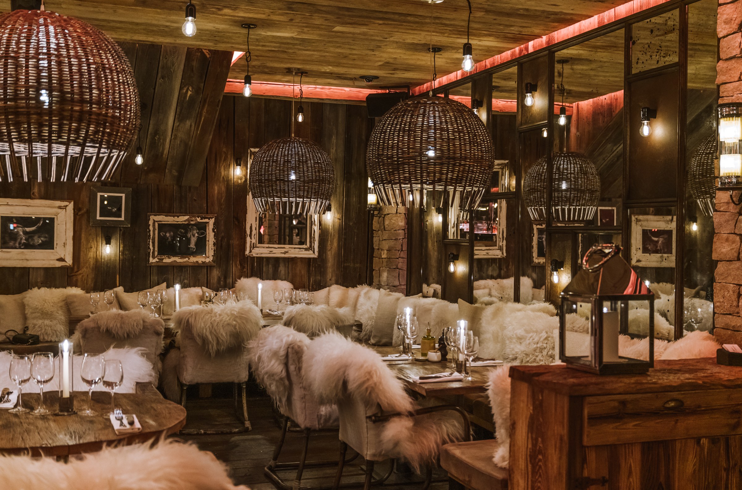 A restaurant interior with fur-covered chairs, ornate wood pendant lights, candles on tables set for dinner, and wood paneling on the walls