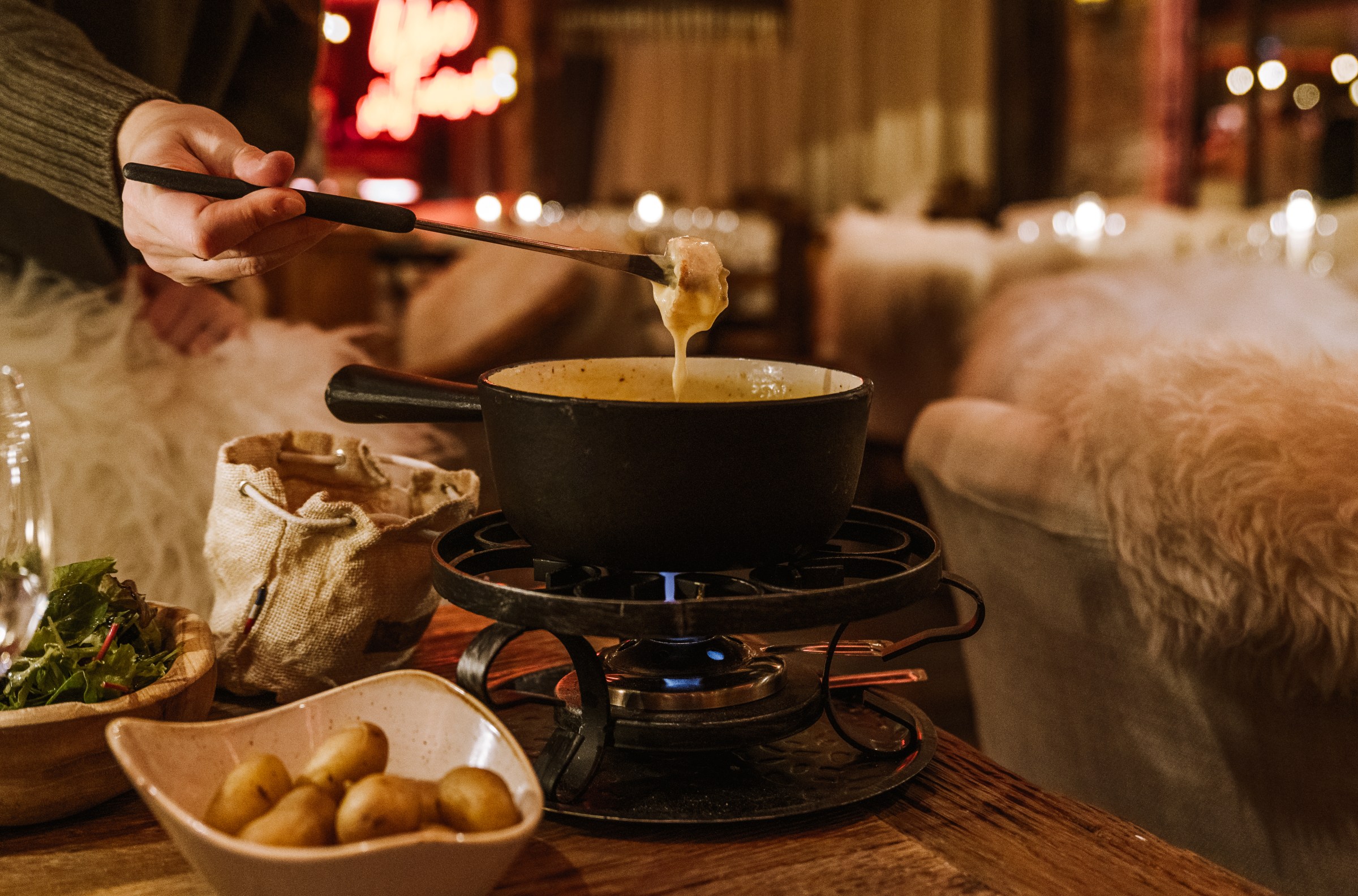 A hand dips a potato into a pot of fondue with a long skewer
