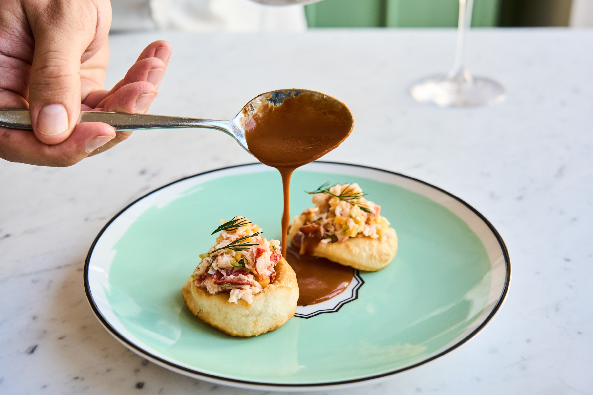 A hand pouring a brown sauce over a lobster-topped pastry.