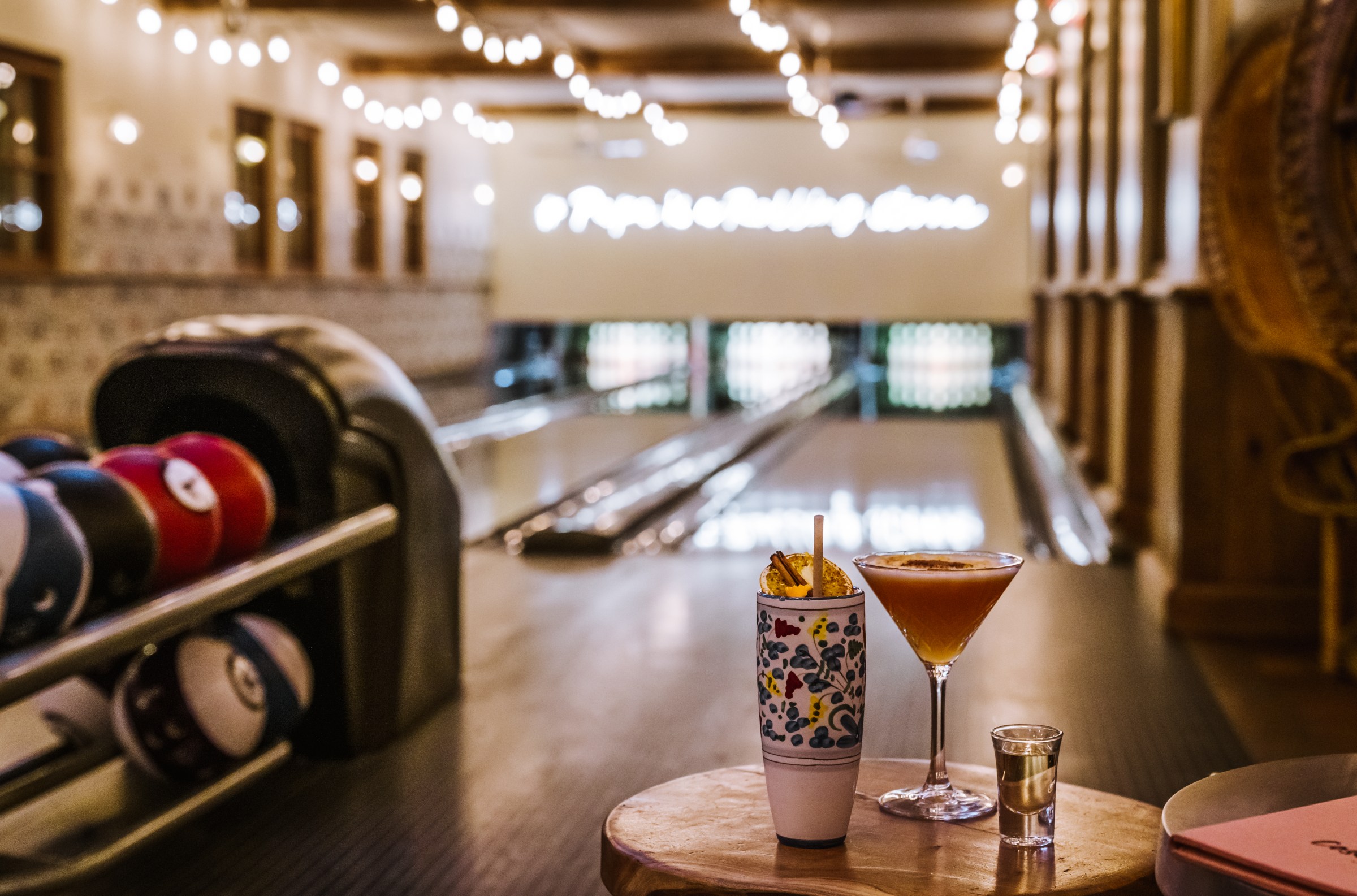 A table with cocktails in front of bowling lanes decorated with a (blurred out) neon sign