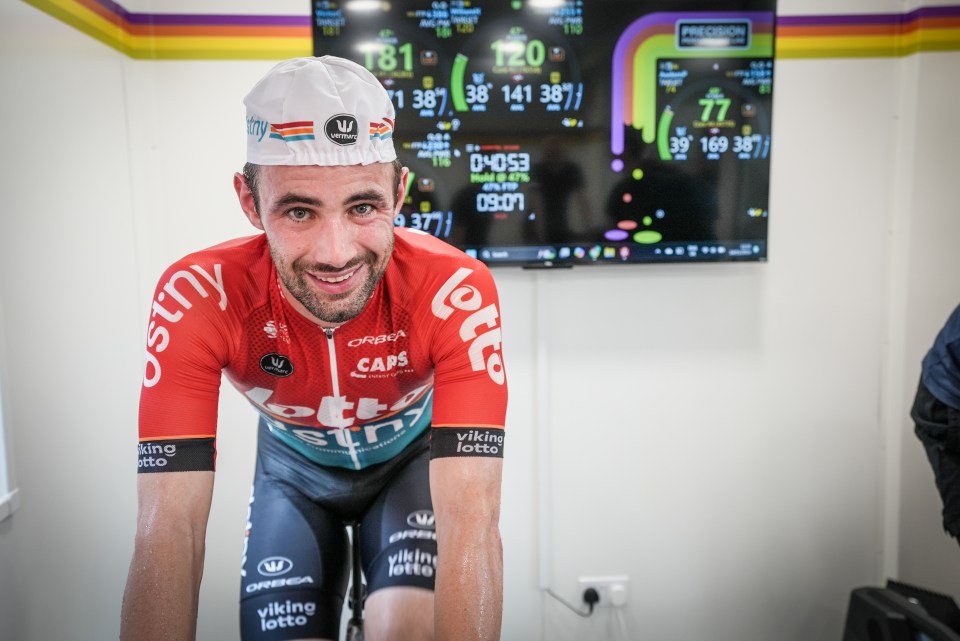 A cyclist in a red and white jersey smiles at the camera while sitting on a stationary bike in front of a performance monitor.