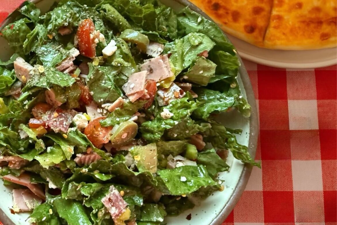 A delicious salad with leafy greens, chopped tomatoes, sliced meat, and cheese sits elegantly on a plate. Hinting at rustic charm, part of a round bread rests nearby on the classic red and white checkered tablecloth—your perfect recipe for a refreshing meal.