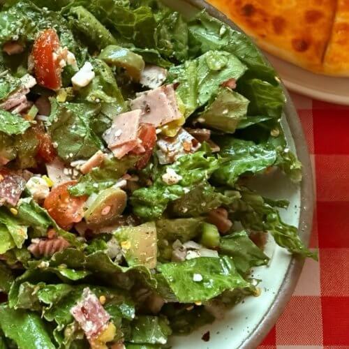 Pizza Shop Salad: The Easy-to-Make Italian Chopped Salad You'll Crave Daily - 3 A delicious salad with leafy greens, chopped tomatoes, sliced meat, and cheese sits elegantly on a plate. Hinting at rustic charm, part of a round bread rests nearby on the classic red and white checkered tablecloth—your perfect recipe for a refreshing meal.