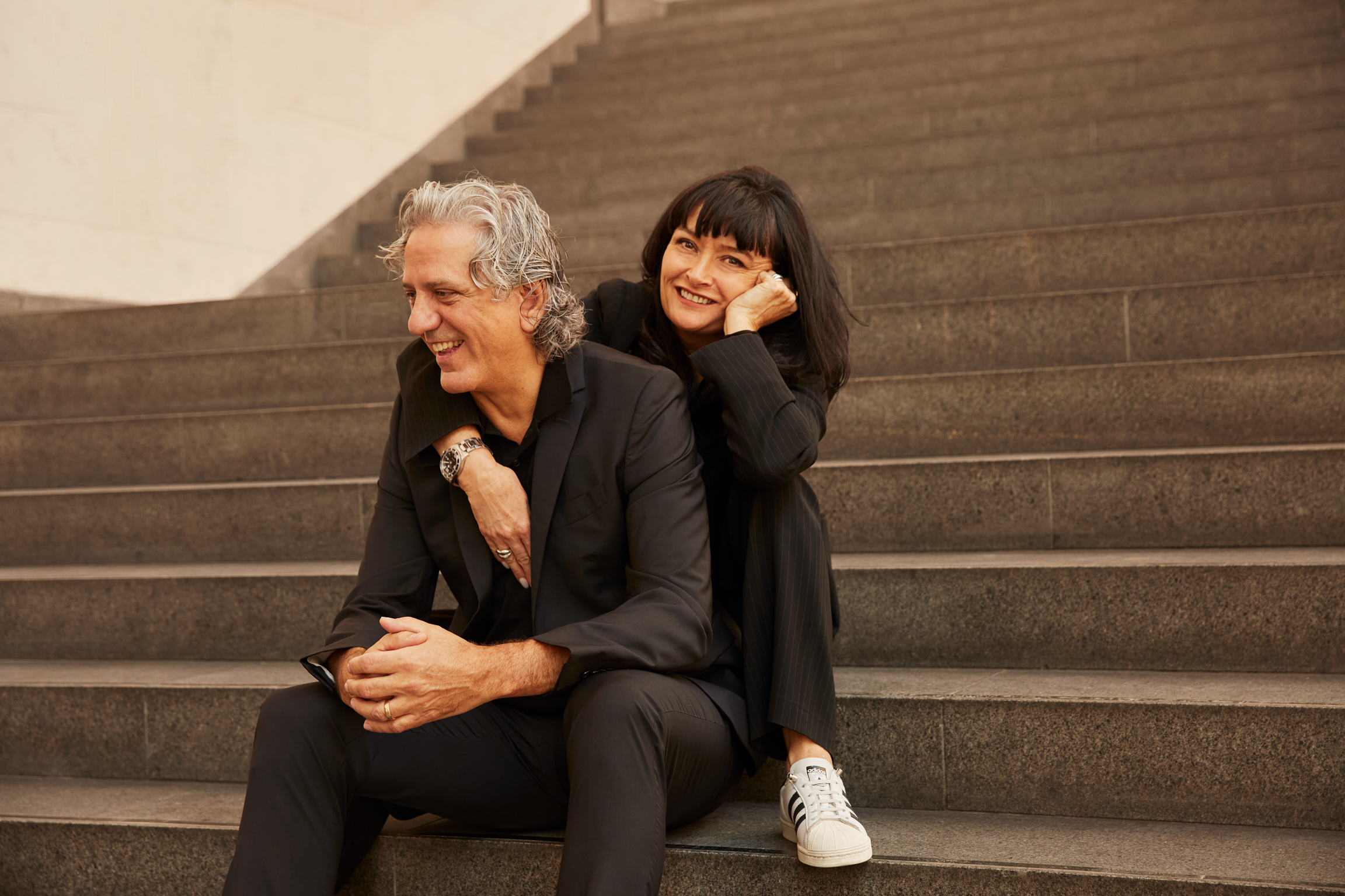 A man with salt and pepper mid-length hair poses for the camera, either sitting at a sleek bronze bar counter, in front of a Caravaggio painting, or sitting on steps while laughing with a woman, dressed in black clothes just like him.