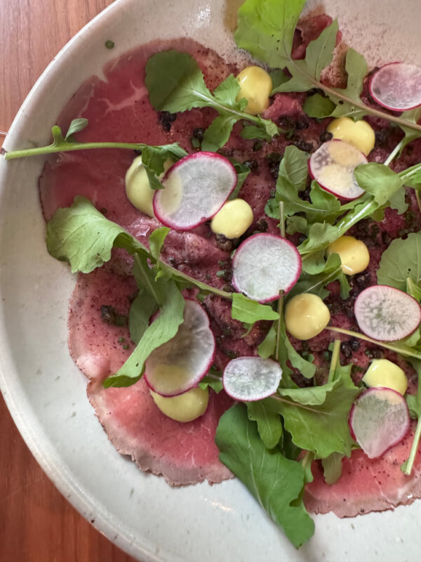 48 Hours in Asheville: A Culinary Tour Through an NC Gem - 12 A plate of beef carpaccio topped with arugula, radish slices, yellow sauce dollops, and greens.