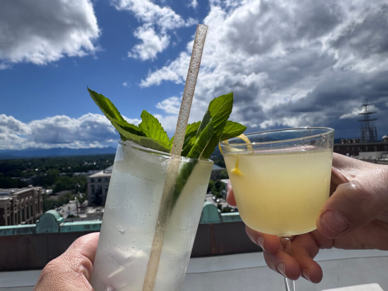 48 Hours in Asheville: A Culinary Tour Through an NC Gem - 11 Two hands holding cocktails—one clear with mint and a straw, the other yellow with a twist—toast above Asheville restaurants on a city rooftop beneath a cloudy sky.