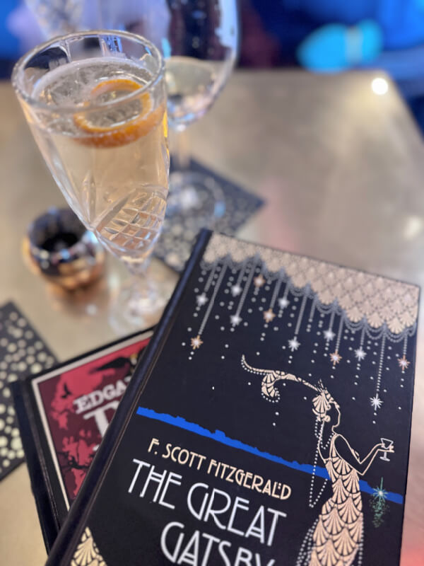 48 Hours in Asheville: A Culinary Tour Through an NC Gem - 3 A glass of cocktail with a slice of orange sits beside a copy of "The Great Gatsby" by F. Scott Fitzgerald on a table, evoking the relaxed charm found in Asheville restaurants.
