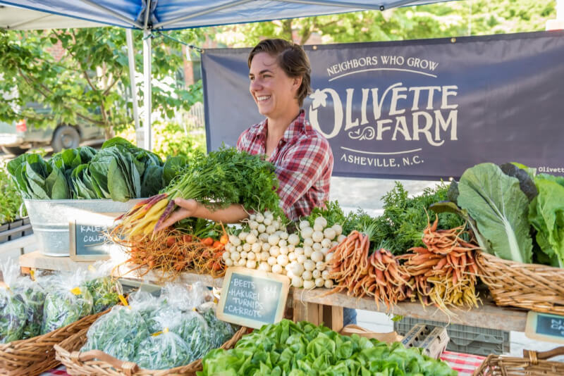 48 Hours in Asheville: A Culinary Tour Through an NC Gem - 14 A vendor smiles behind a table of fresh vegetables, including carrots, beets, and greens, at an outdoor Olivette Farm stand.