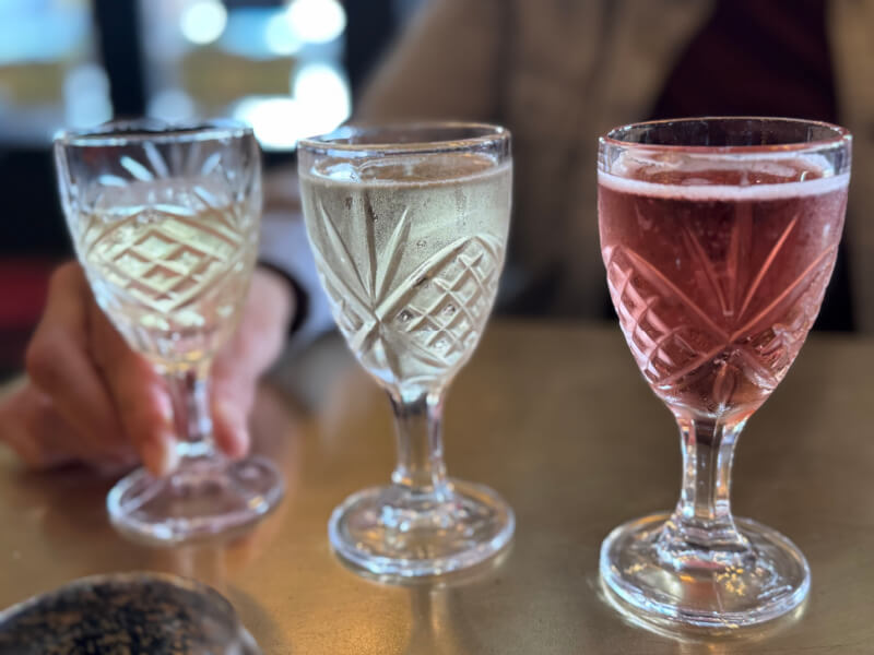 48 Hours in Asheville: A Culinary Tour Through an NC Gem - 2 Three crystal glasses containing different colored beverages—white, clear, and pink—are placed on a table, with a hand holding the glass on the left.