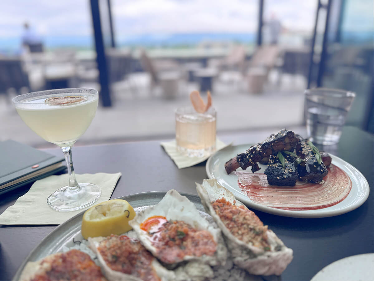 48 Hours in Asheville: A Culinary Tour Through an NC Gem - 1 A table at an outdoor Asheville restaurant features a cocktail, a short drink, a plate of seasoned oysters with lemon, a serving of ribs with garnish, and a glass of water.