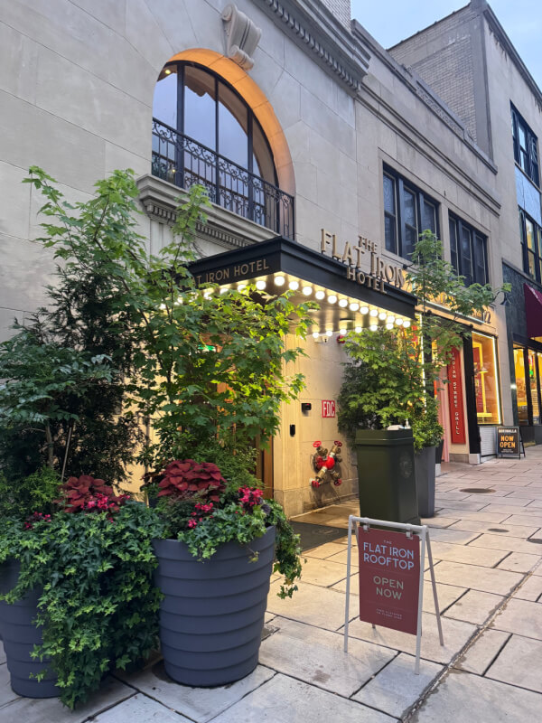 48 Hours in Asheville: A Culinary Tour Through an NC Gem - 10 Entrance of The Flat Iron Hotel with marquee lights, potted plants, and a sign advertising the rooftop. The sidewalk and neighboring storefronts are visible.