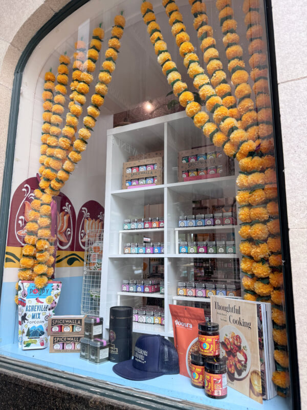 48 Hours in Asheville: A Culinary Tour Through an NC Gem - 17 A shop window display featuring colorful spice jars, cookbooks, canned goods, a navy hat, and yellow flower garlands hanging along the glass.