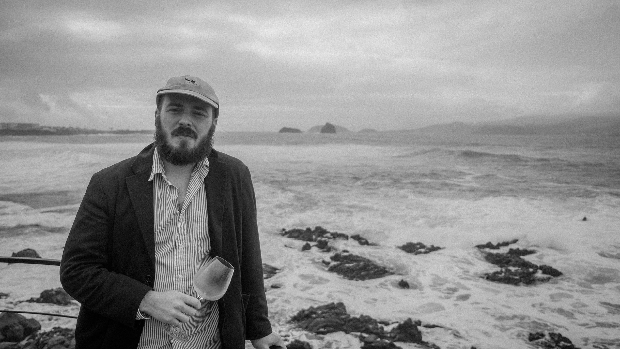 Black and white headshot of Lewis Kopman who poses with an empty wine glass in front of a large body of water.