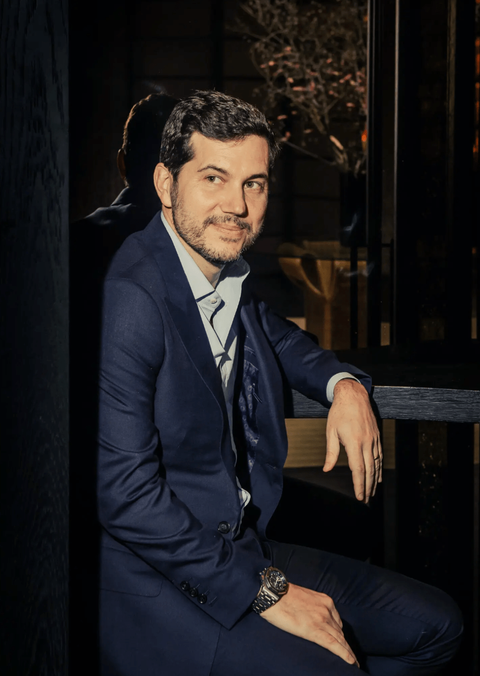  A professional portrait of Scott Sartiano dressed in a sharp suit, seated and smiling confidently, with an elegant restaurant background.