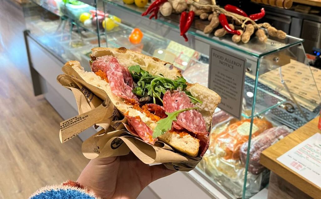 ad maiora sandwich in hand in manchester sandwich shop with deli counter in background