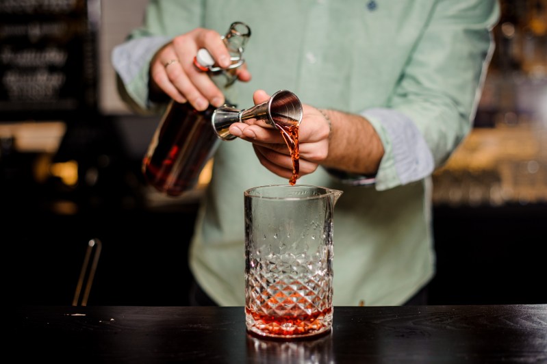 Bartender making alcoholic cocktail in red color, metal jigger and bar environment