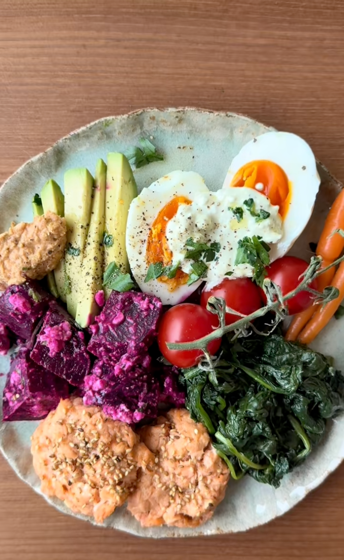 A vibrant plate featuring avocado slices, boiled eggs, cherry tomatoes, cooked greens, beet salad, carrot sticks, and hummus, arranged artfully