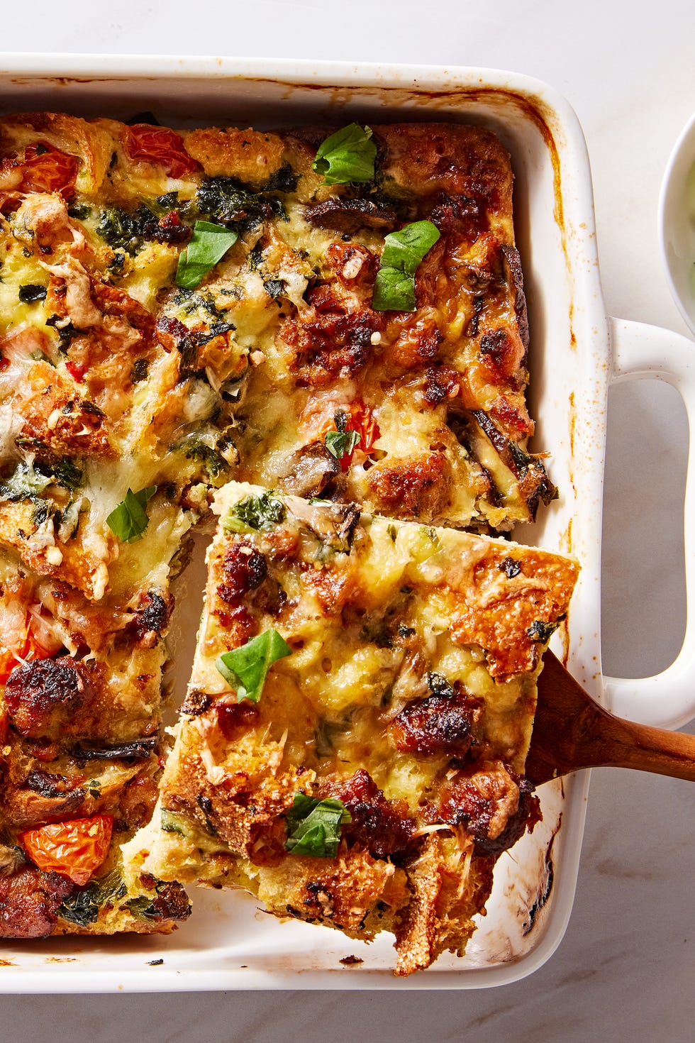 eggy breakfast strata featuring sausage, tomatoes, bread, and mushrooms, and topped with basil