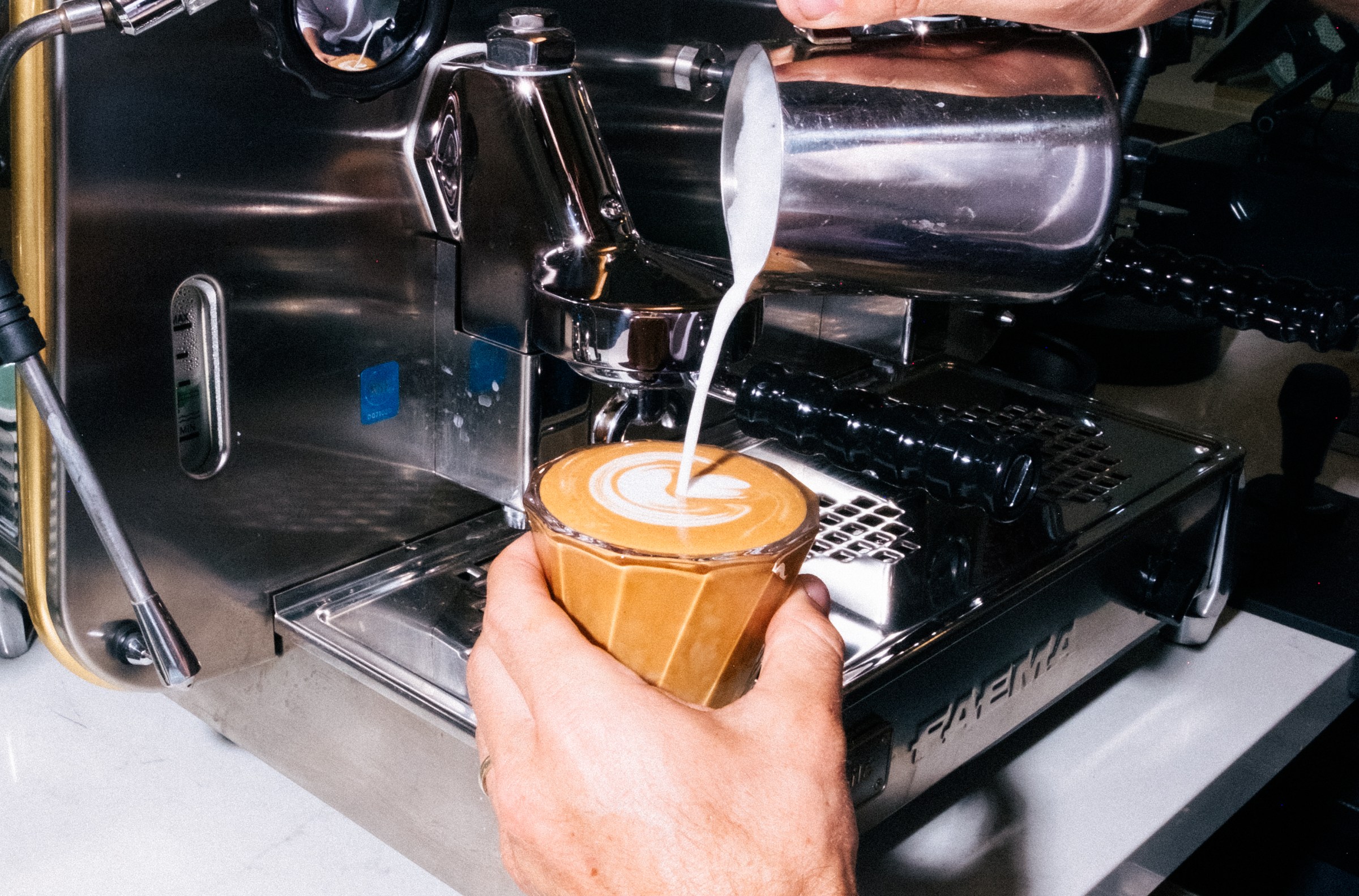 Hand pouring steamed milk into a latte in a clear glass cup