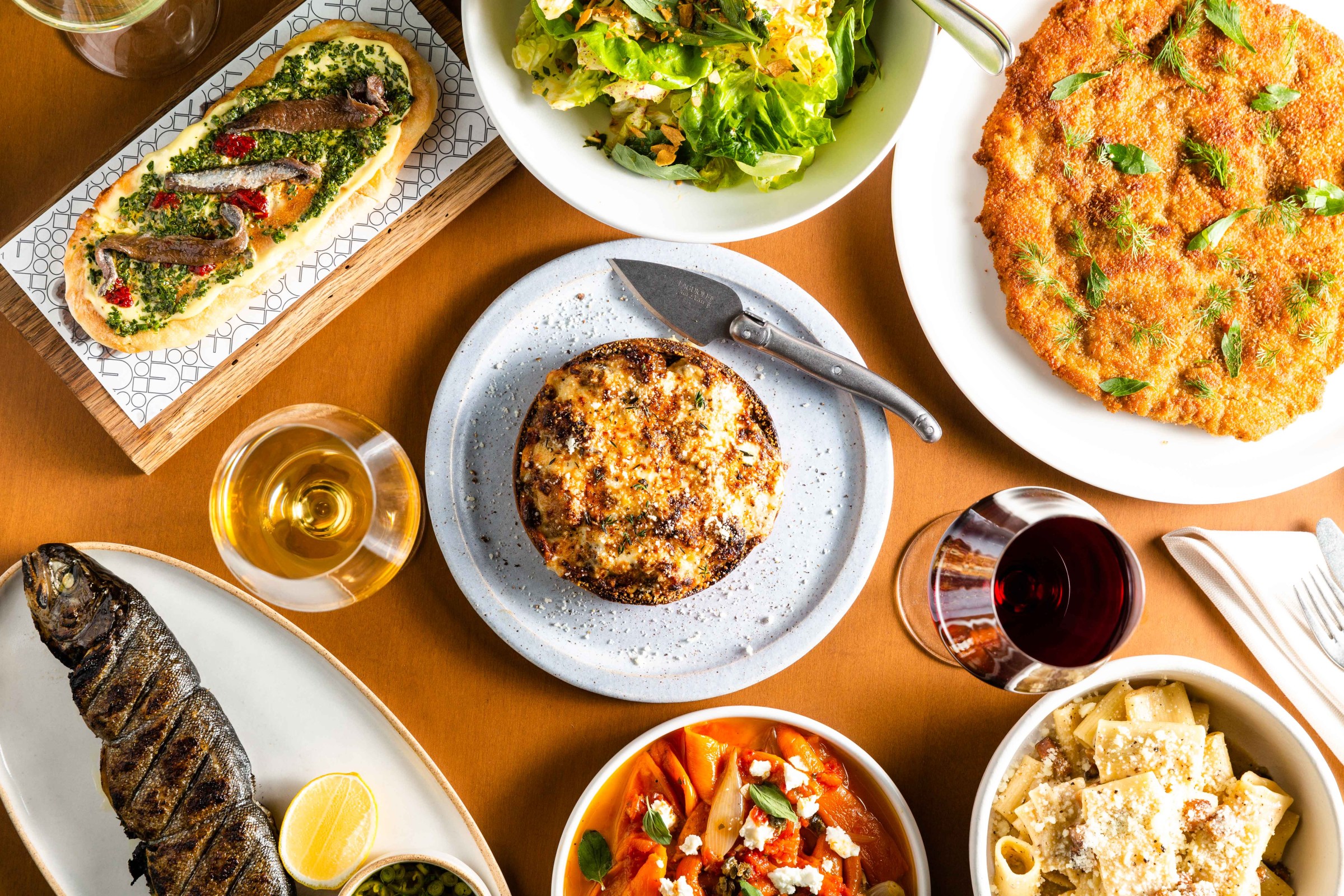 A spread of dishes from Ci Siamo’s menu laid out on a light wooden table interspersed with two glasses of wine.