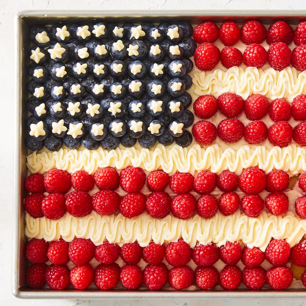 white cake decorated to look like an american flag with blueberries for stars, raspberries for red stripes, and off white frosting for white stripes