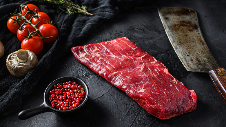 A raw bavette steak (or flap steak) on a black stone surface, surrounded by a butcher knife, a tomatoes on the vine, a mushroom, a sprig of rosemary, and a dish of red peppercorns.