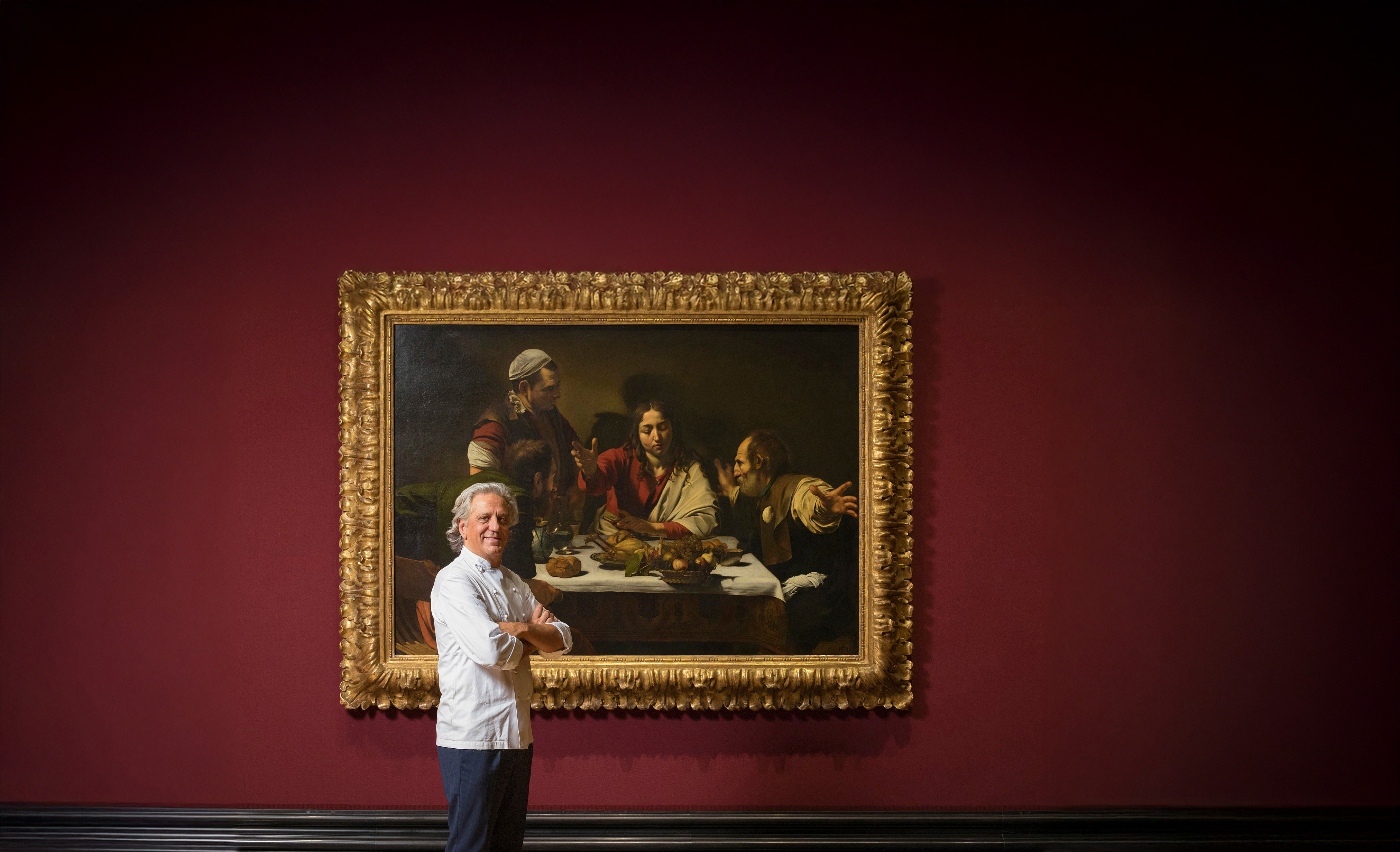 A man with salt and pepper mid-length hair poses for the camera, either sitting at a sleek bronze bar counter, in front of a Caravaggio painting, or sitting on steps while laughing with a woman, dressed in black clothes just like him.