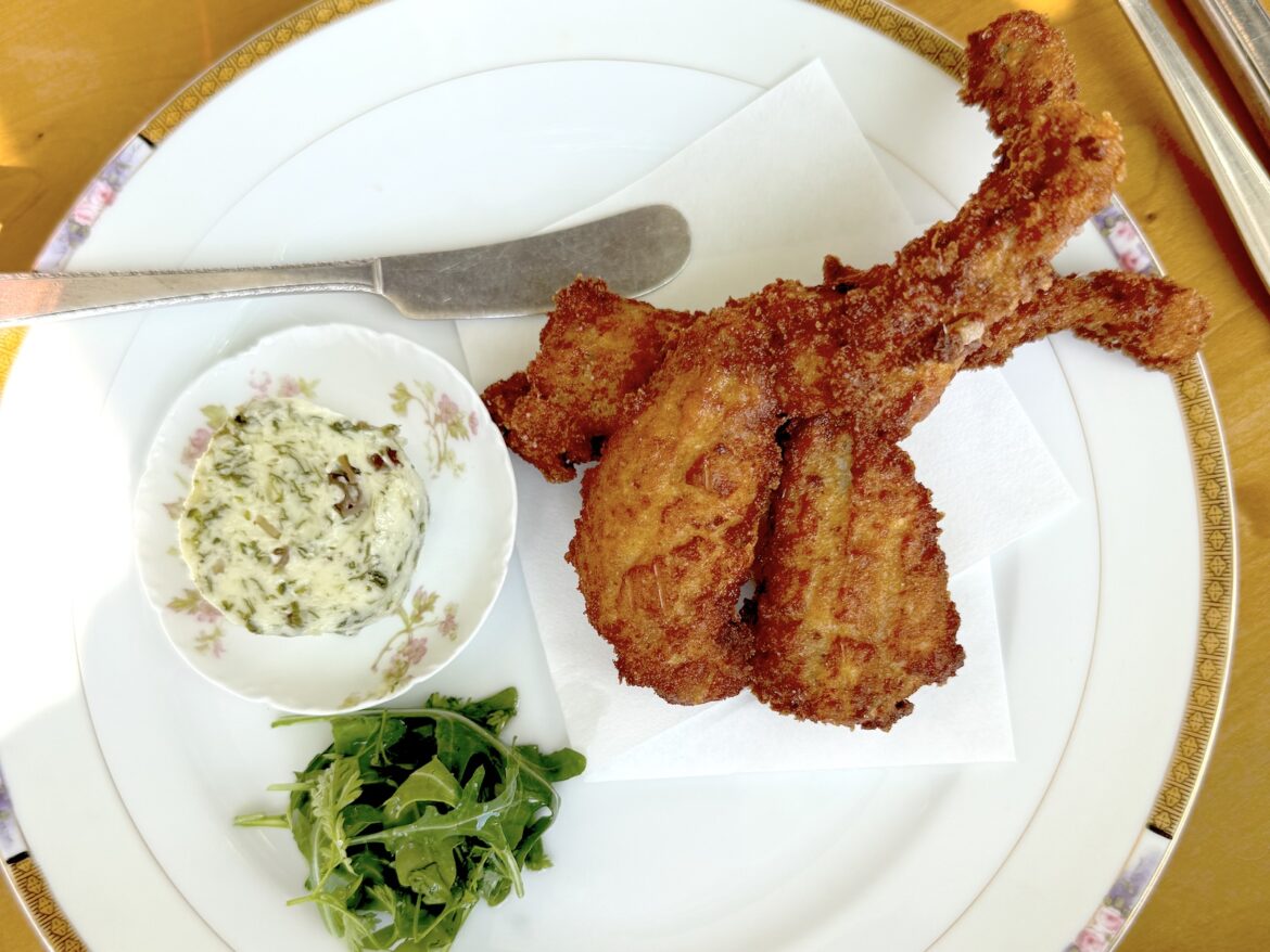 Why Can’t French Cuisine Make it in Denver? Three deep-fried frog legs on a plate with fresh greens, a small bowl of compound butter, and a butter knife