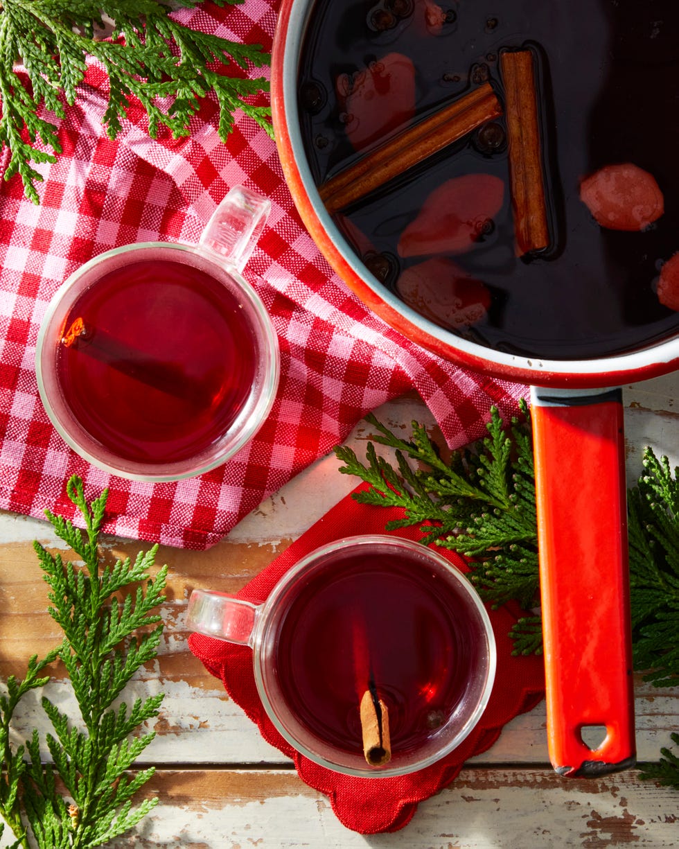 two clear cups of dark red liquid beside a pot of the same liquid with cinnamon sticks and greenery
