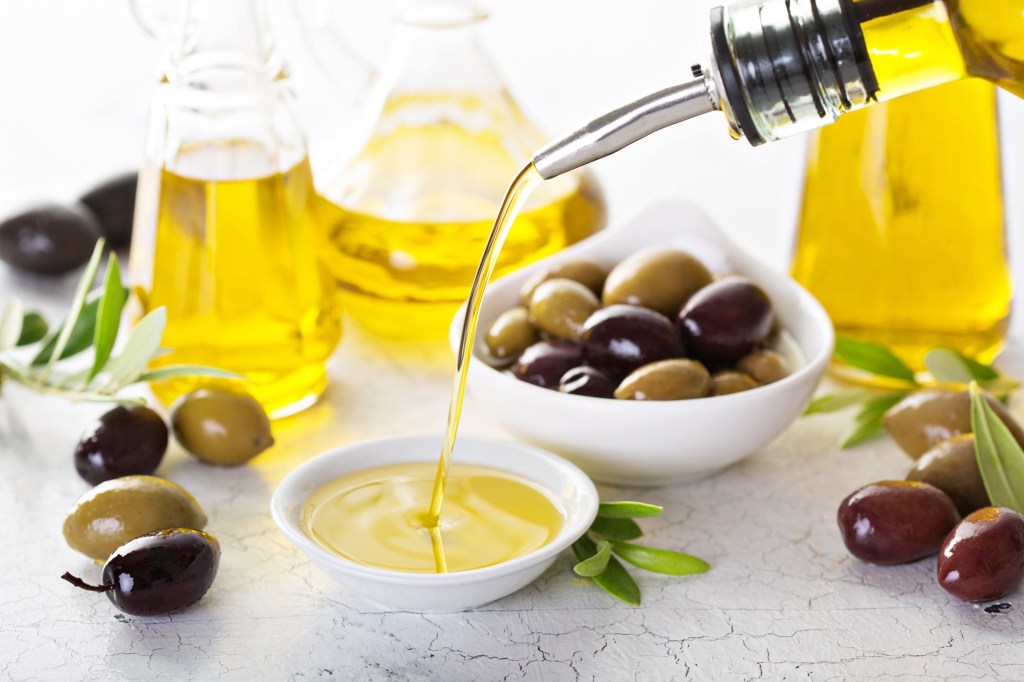 Olive oil being poured into a small bowl next to olives and olive oil bottles.