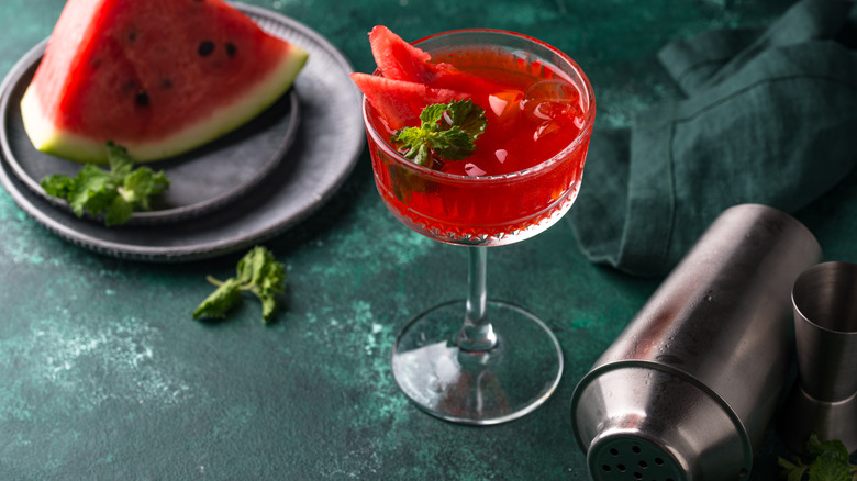 Slice of watermelon on a plate with a dark red cocktail and shaker