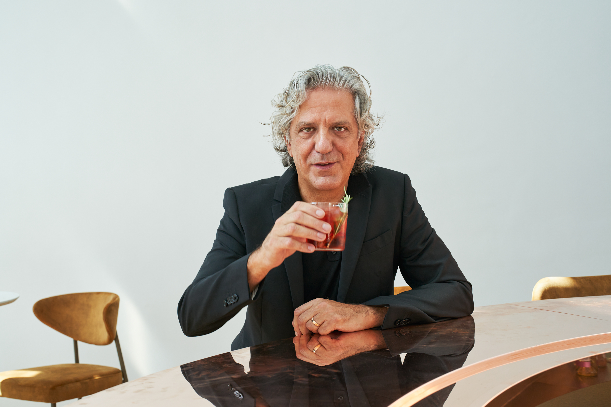 A man with salt and pepper mid-length hair poses for the camera, either sitting at a sleek bronze bar counter, in front of a Caravaggio painting, or sitting on steps while laughing with a woman, dressed in black clothes just like him.