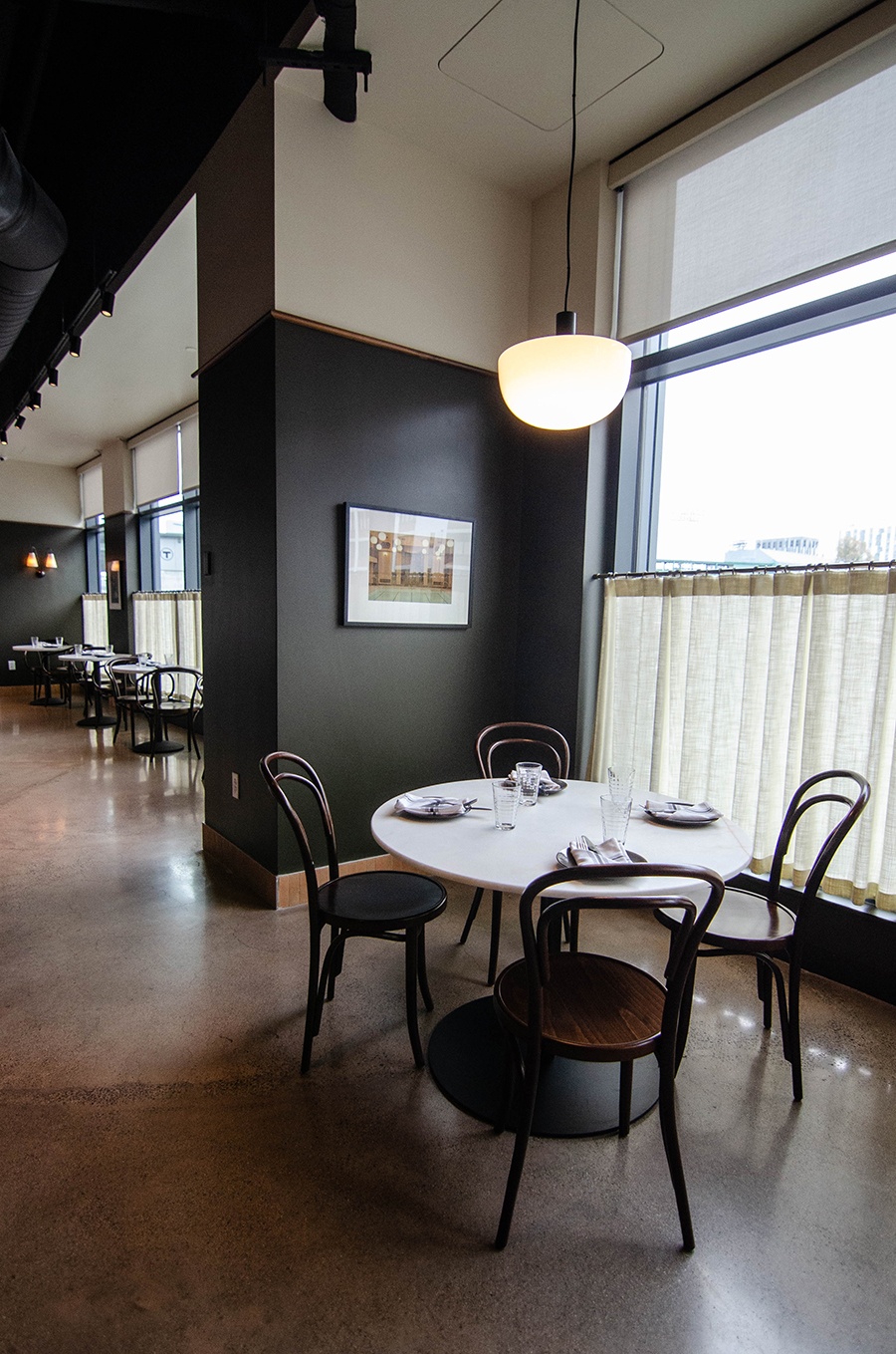 A restaurant dining room features a small white table with four wooden chairs by a window with cafe curtains.