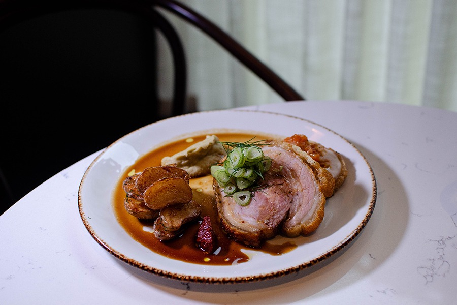 Porchetta with crispy skin and roasted fingerling potatoes sit on a plate in a thin brown sauce.