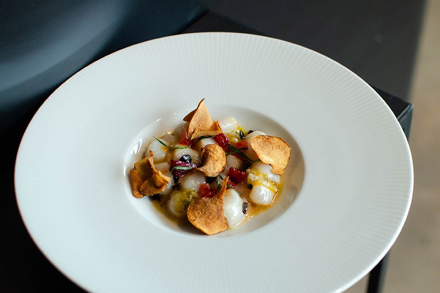 A raw scallop crudo sits in an orange sauce with small sunchoke chips as garnish.