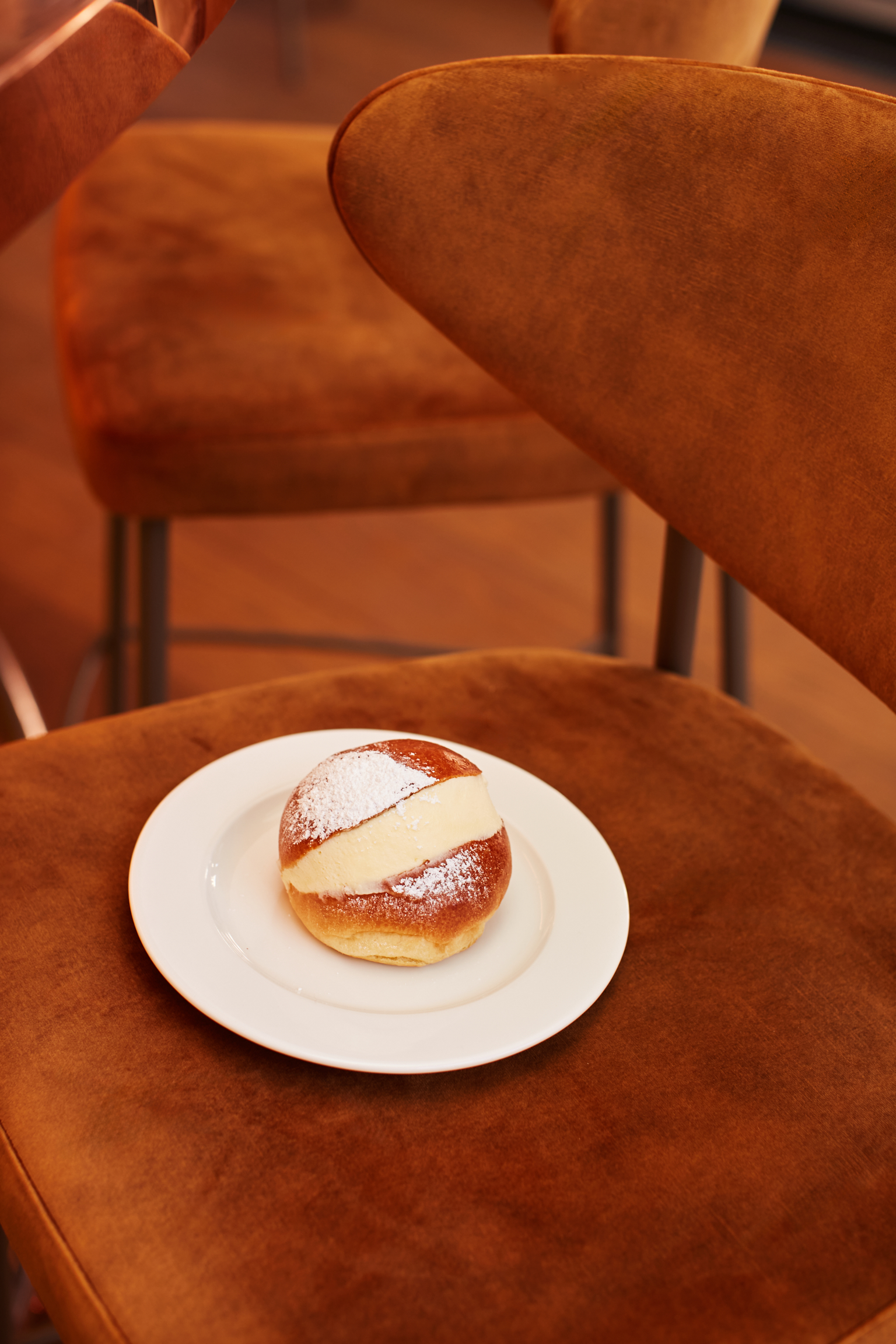 A maritozzo cream-filled bun sits in a white plate placed atop a brown velvet chair.