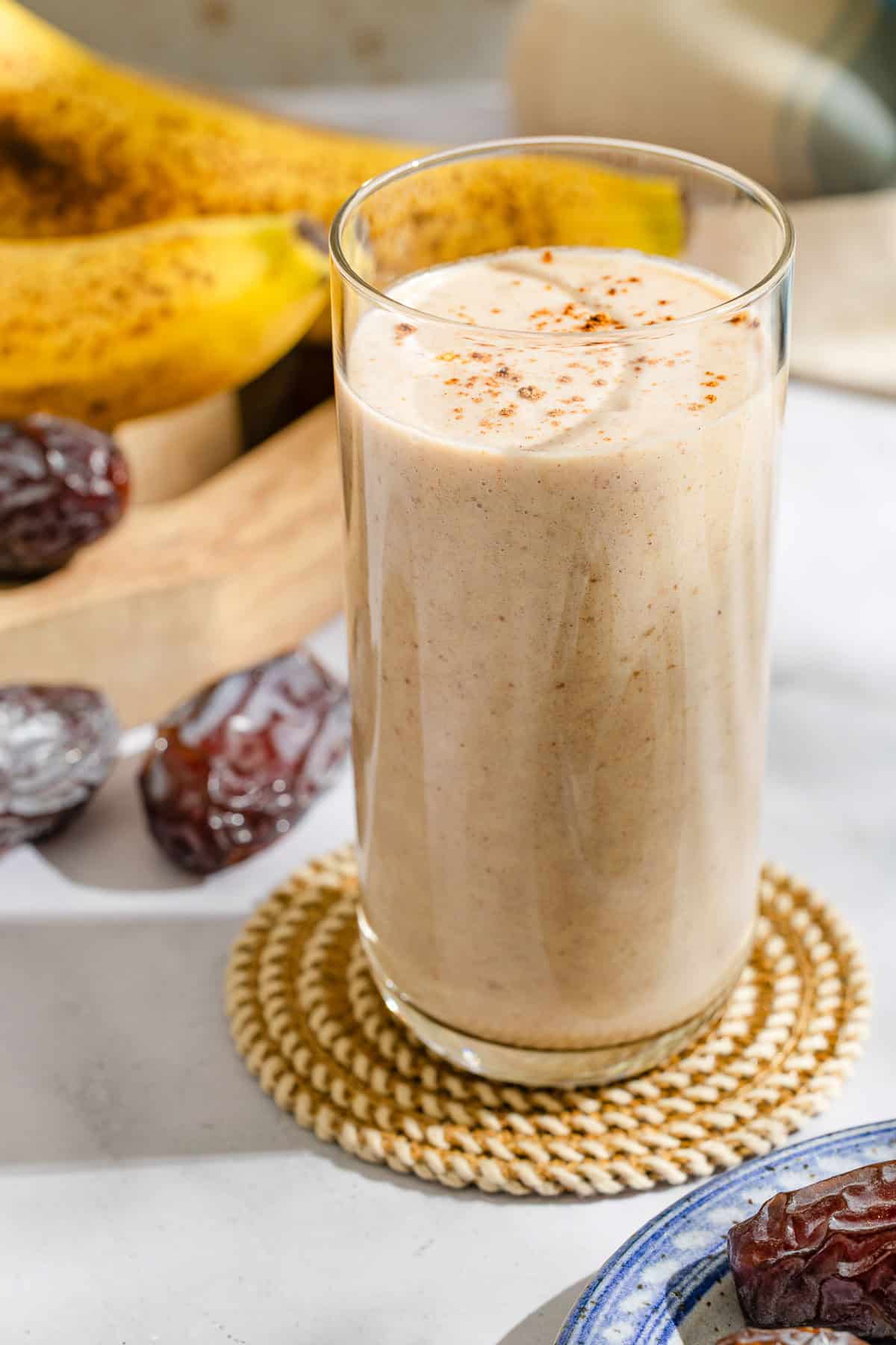 A date shake in a glass on a coaster surrounded by bananas, dates and a kitchen towel.