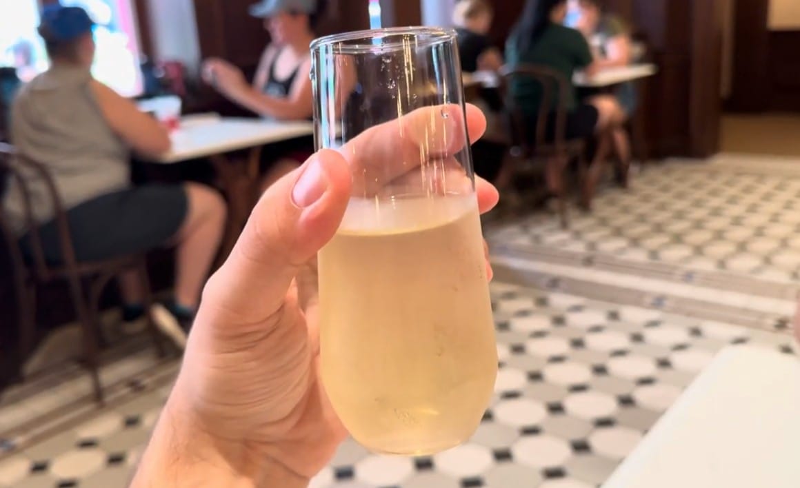 A hand holds a light drink at Cafe L’air de la Sirène, with people enjoying the Epic Universe vibe in the background.