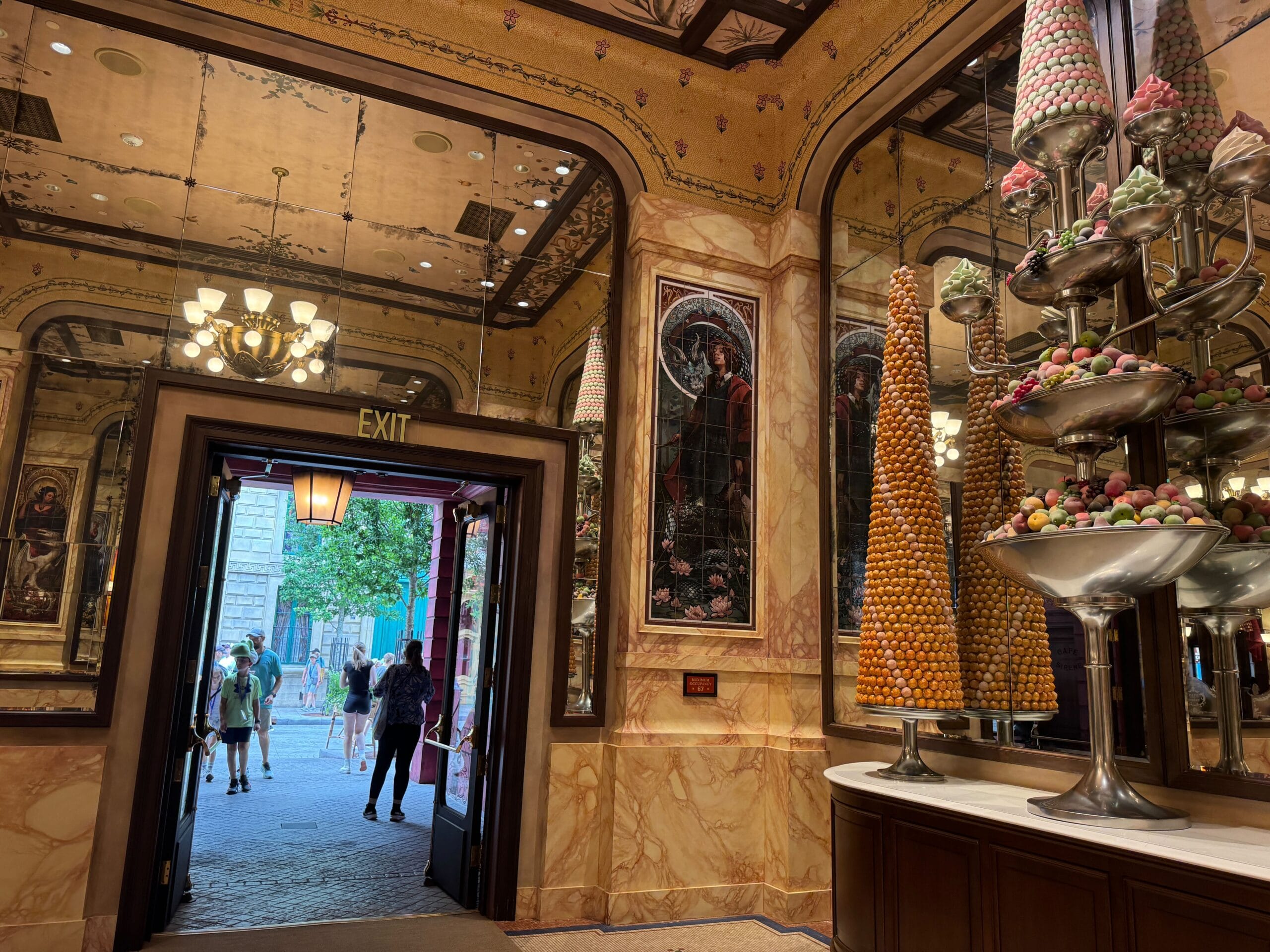 Ornate Cafe L’air de la Sirène pastry shop with tall dessert towers; open door reveals Epic Universe visitors outside.