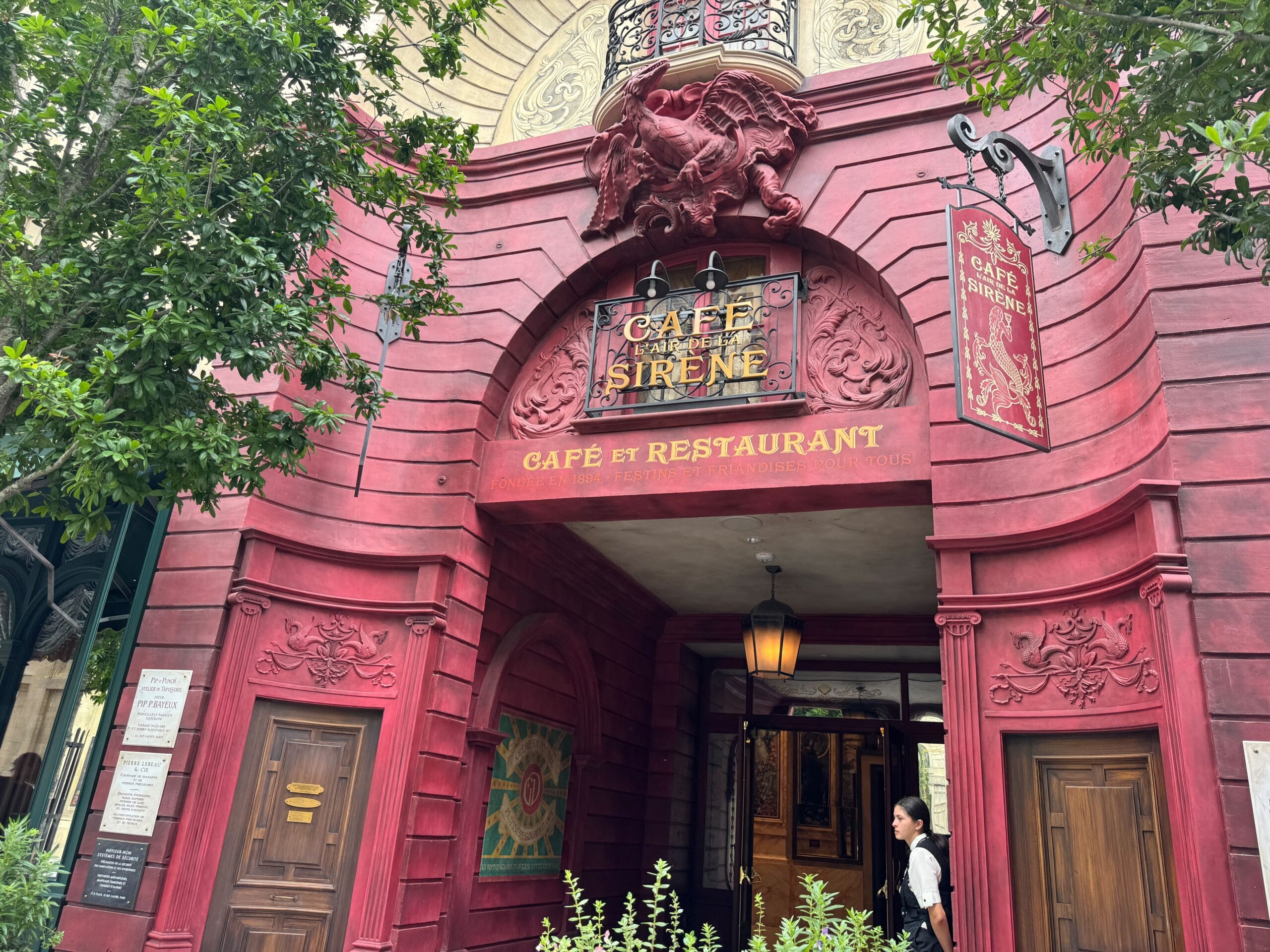 A red Cafe L’air de la Sirène entrance with ornate decor, a hanging sign, and someone at the doorway.
